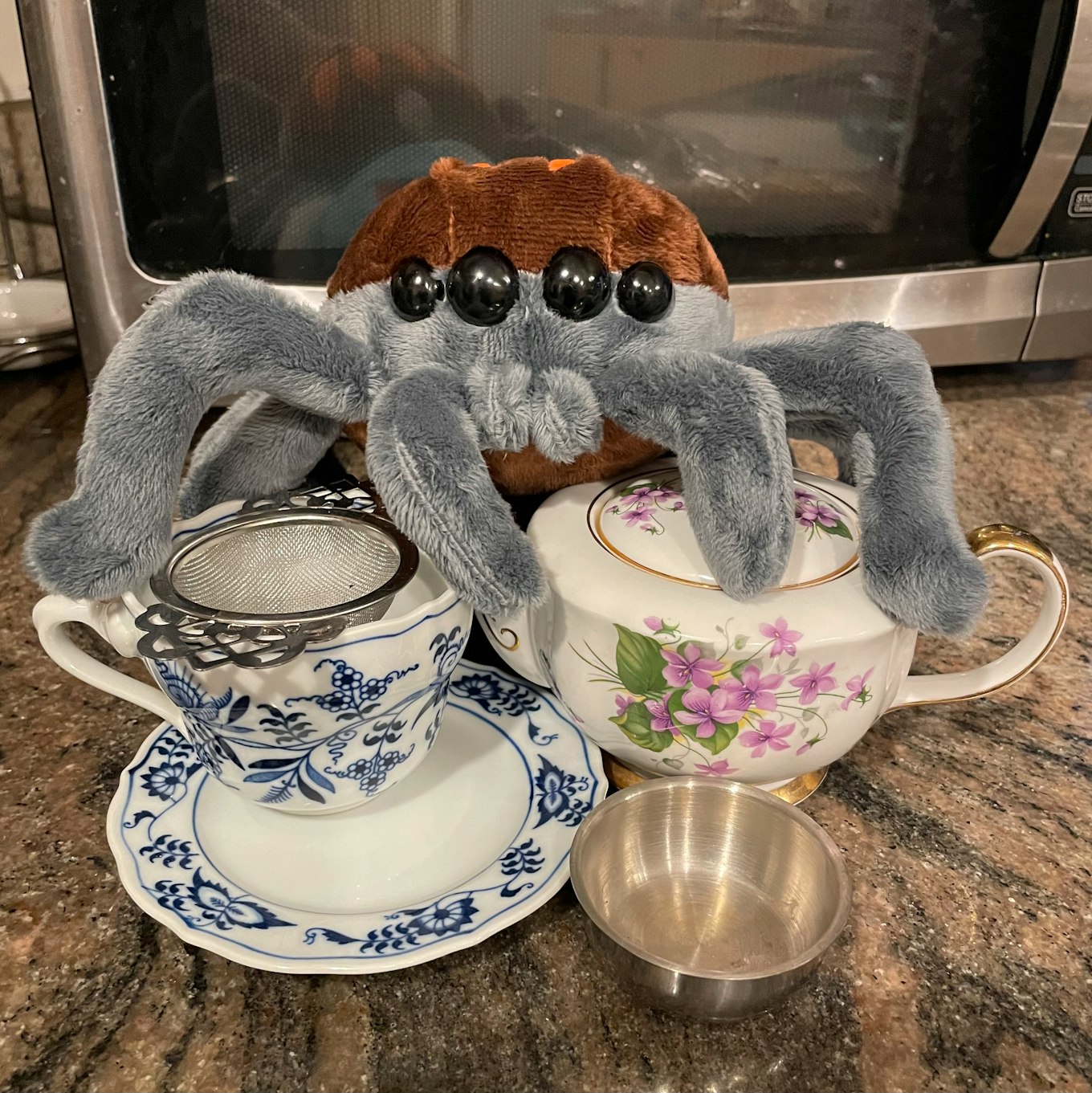 A plush spider sitting over a tea cup and teapot 